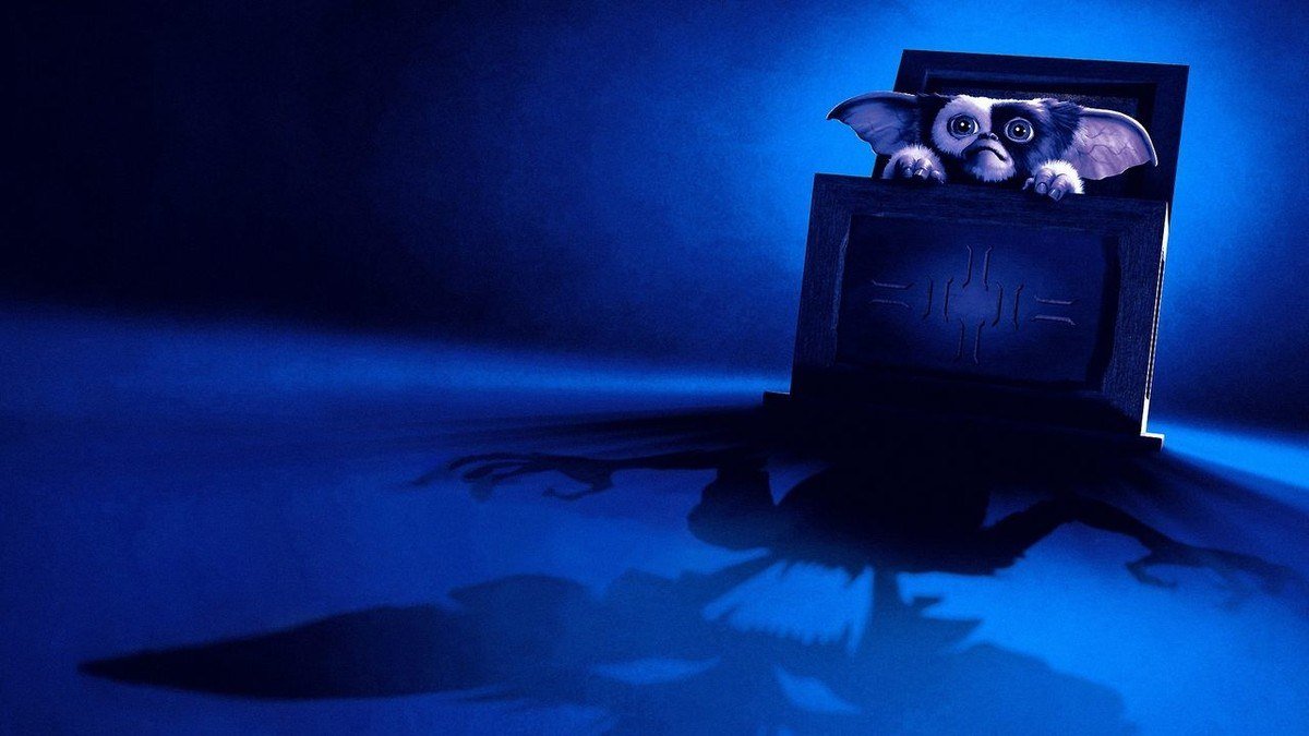 Gremlins key art with Gizmo peeking out of a wooden box while a gremlin shadow spreads below
