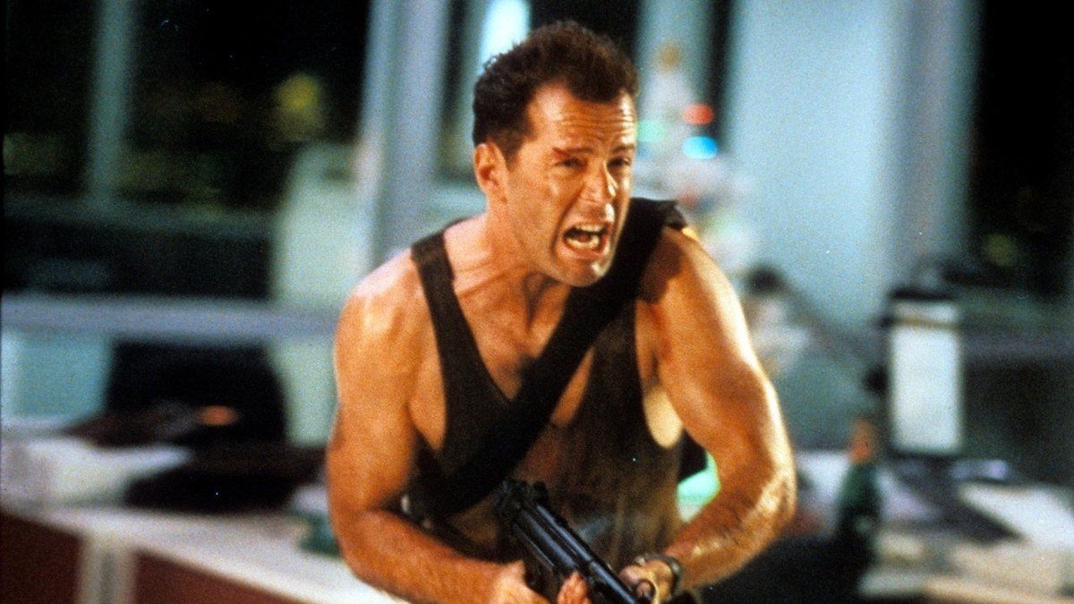 Still from Die Hard with Bruce Willis as John McClane in a sweat-stained vest holding a rifle
