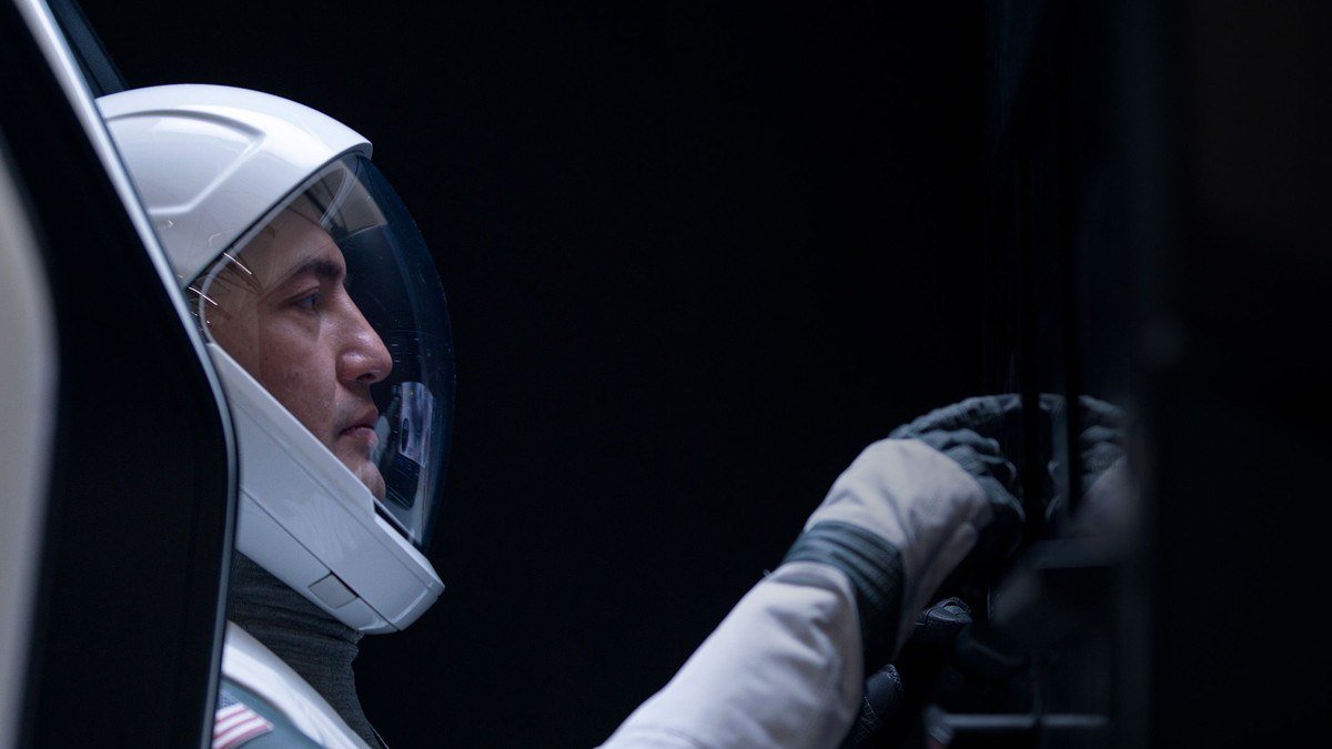 Astronaut in a white Dragon flight suit reaching toward controls in a dark cabin