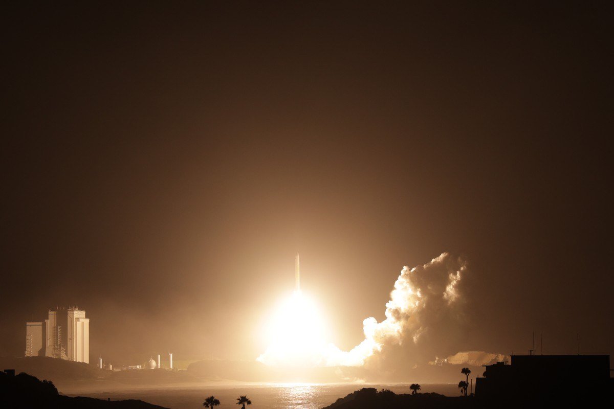 A rocket lifting off at night with a bright flame and a plume of smoke beside a launch complex