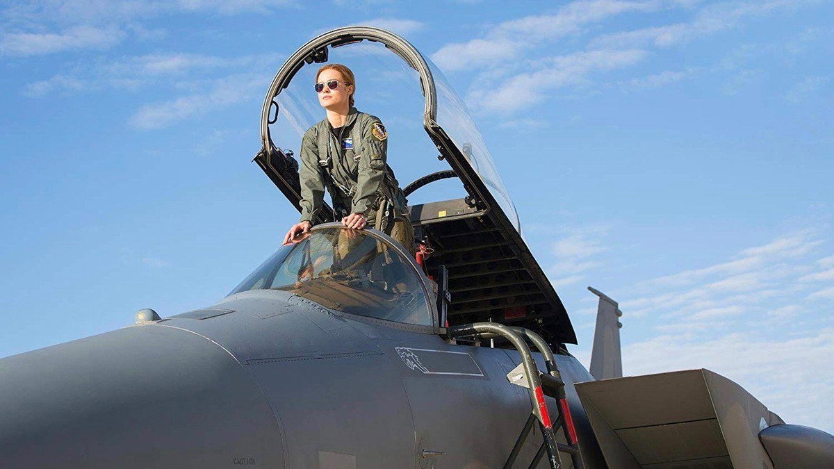 Carol Danvers stands in the cockpit of a fighter jet on a sunny airfield