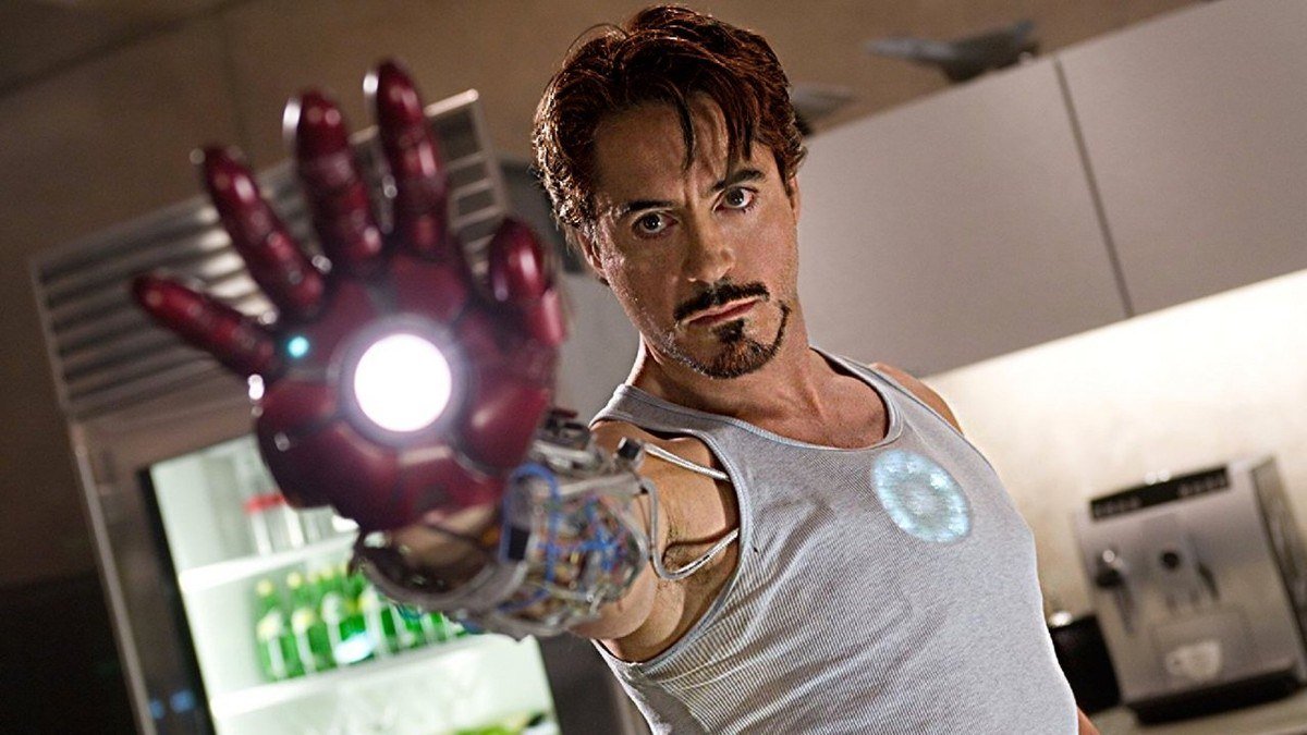 Tony Stark in a tank top raises his repulsor glove toward the camera in his kitchen