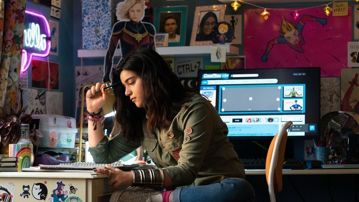 Kamala Khan sketches at a desk in a bedroom covered with Captain Marvel posters
