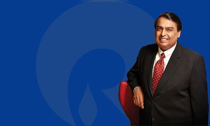 Mukesh Ambani in a dark suit with a red tie standing beside a red chair against a blue Reliance-logo backdrop