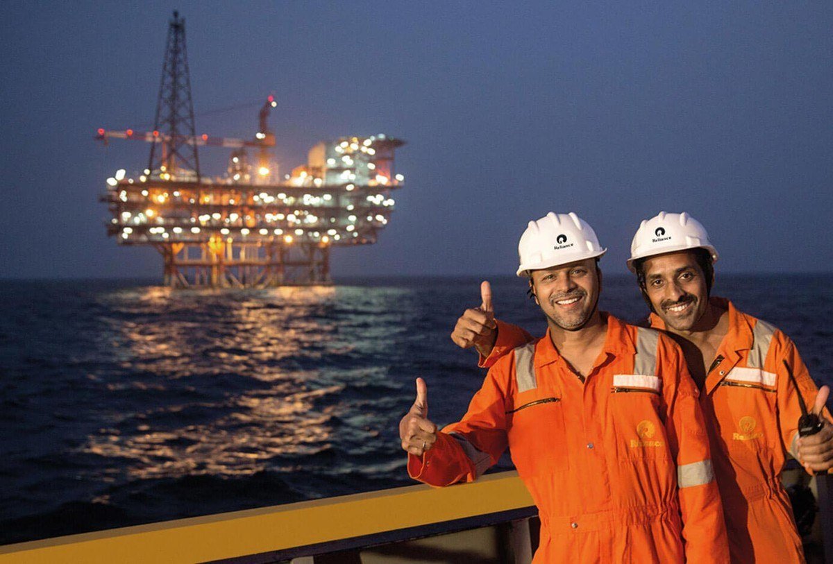 Two smiling offshore workers in orange coveralls and white helmets with a lit platform behind them at dusk